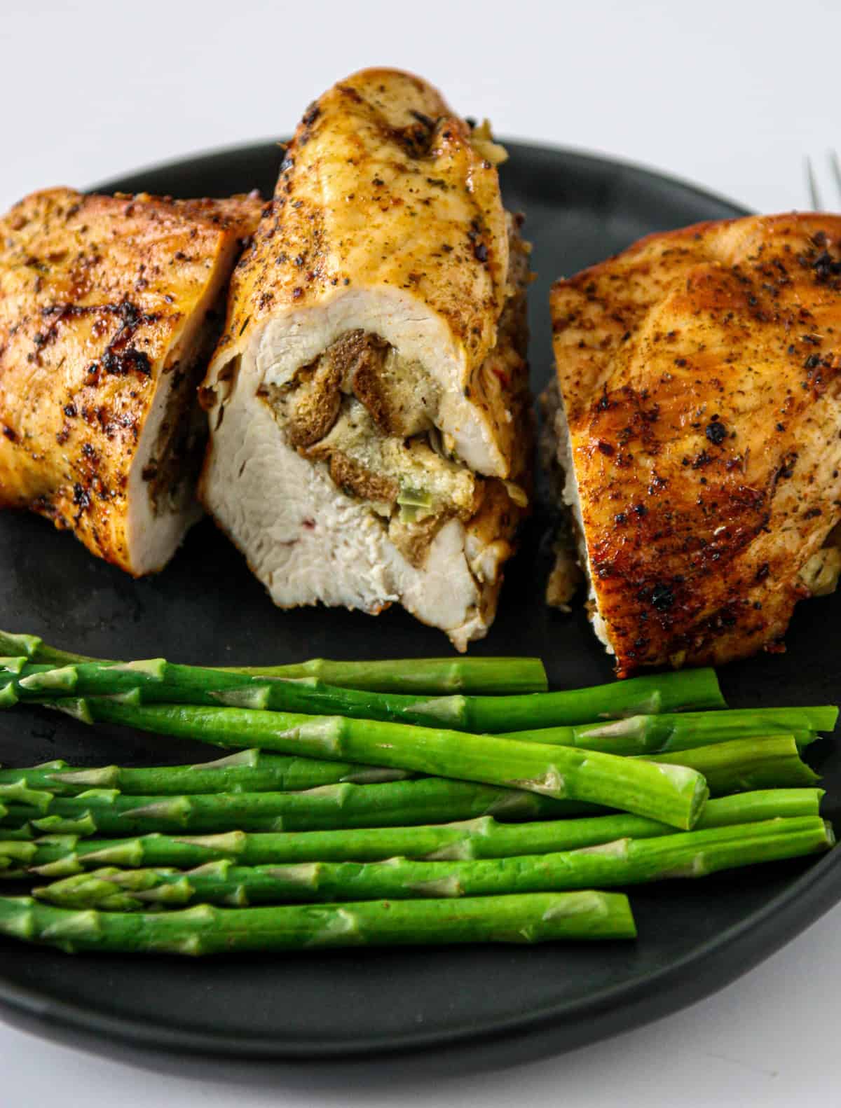 Three stuffing stuffed chicken breast halves on a black plate with asparagus.